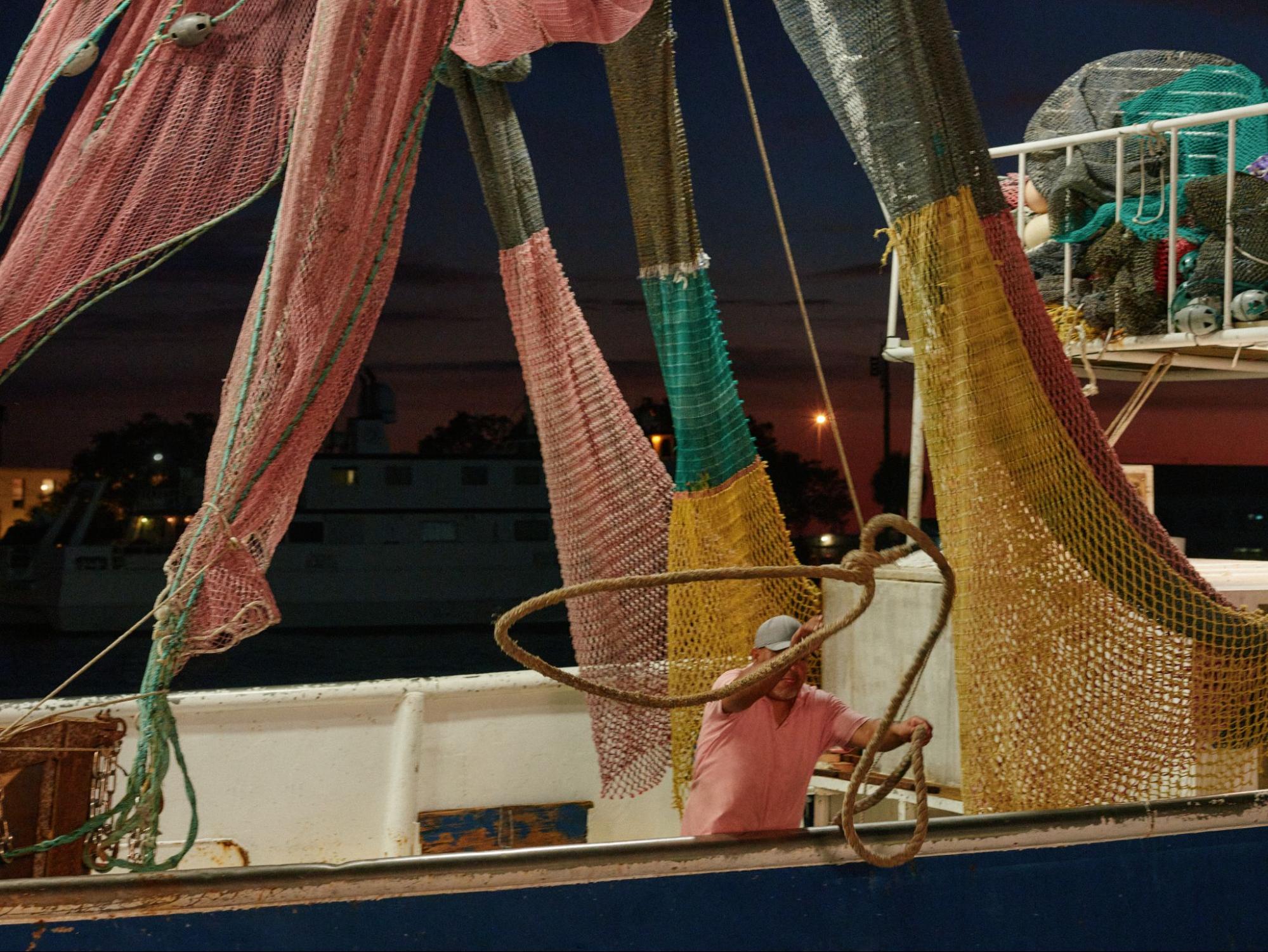 A fisherman in a pink shirt coils rope on the deck of a shrimp boat beneath hanging nets at dusk.