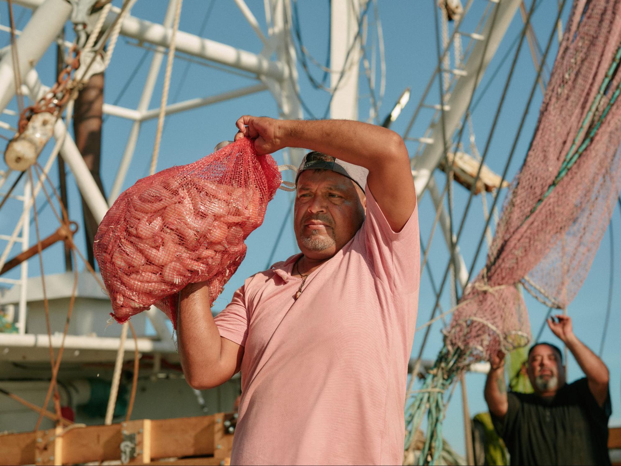 A fisherman in a pink shirt hoists a red mesh bag of shrimp on the deck of a boat.