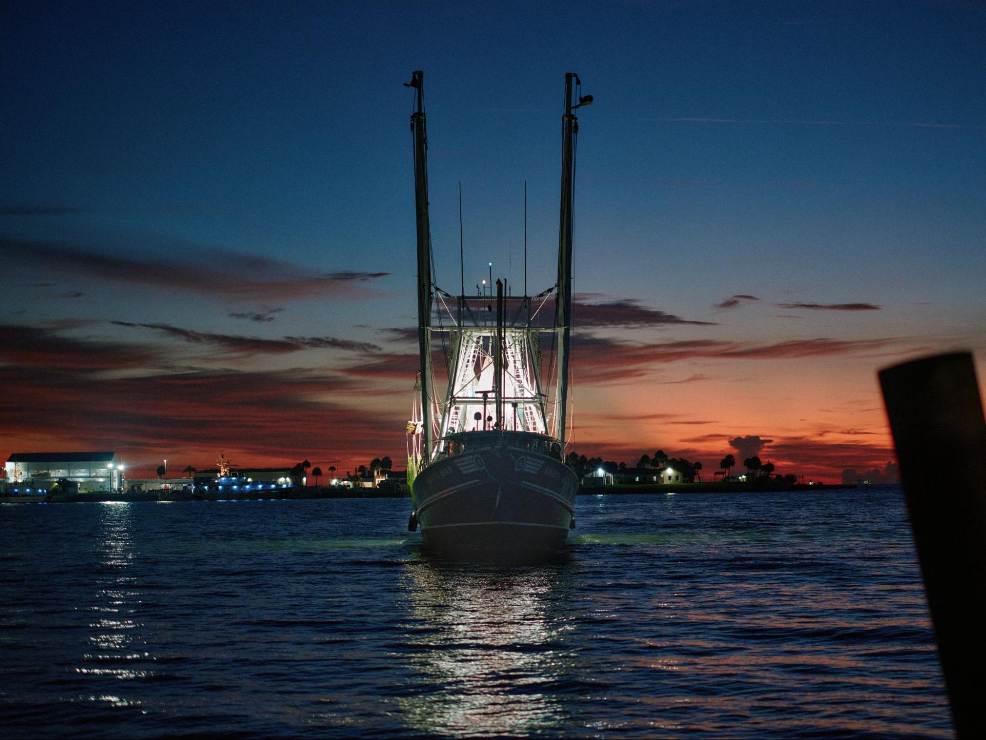 A shrimp boat, its rigging lit from within, on dark water at sunset.