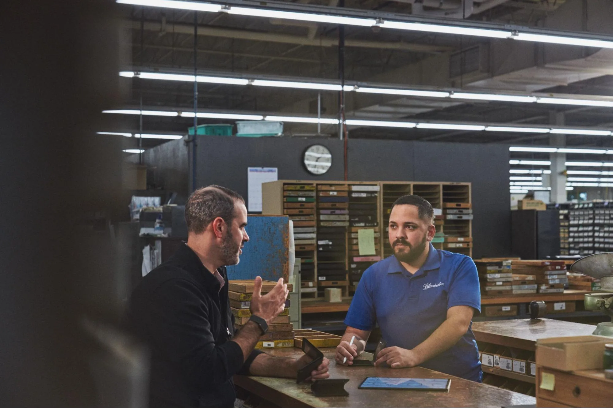 Two Blackinton team members discuss an order at a workbench surrounded by trays of badge dies.