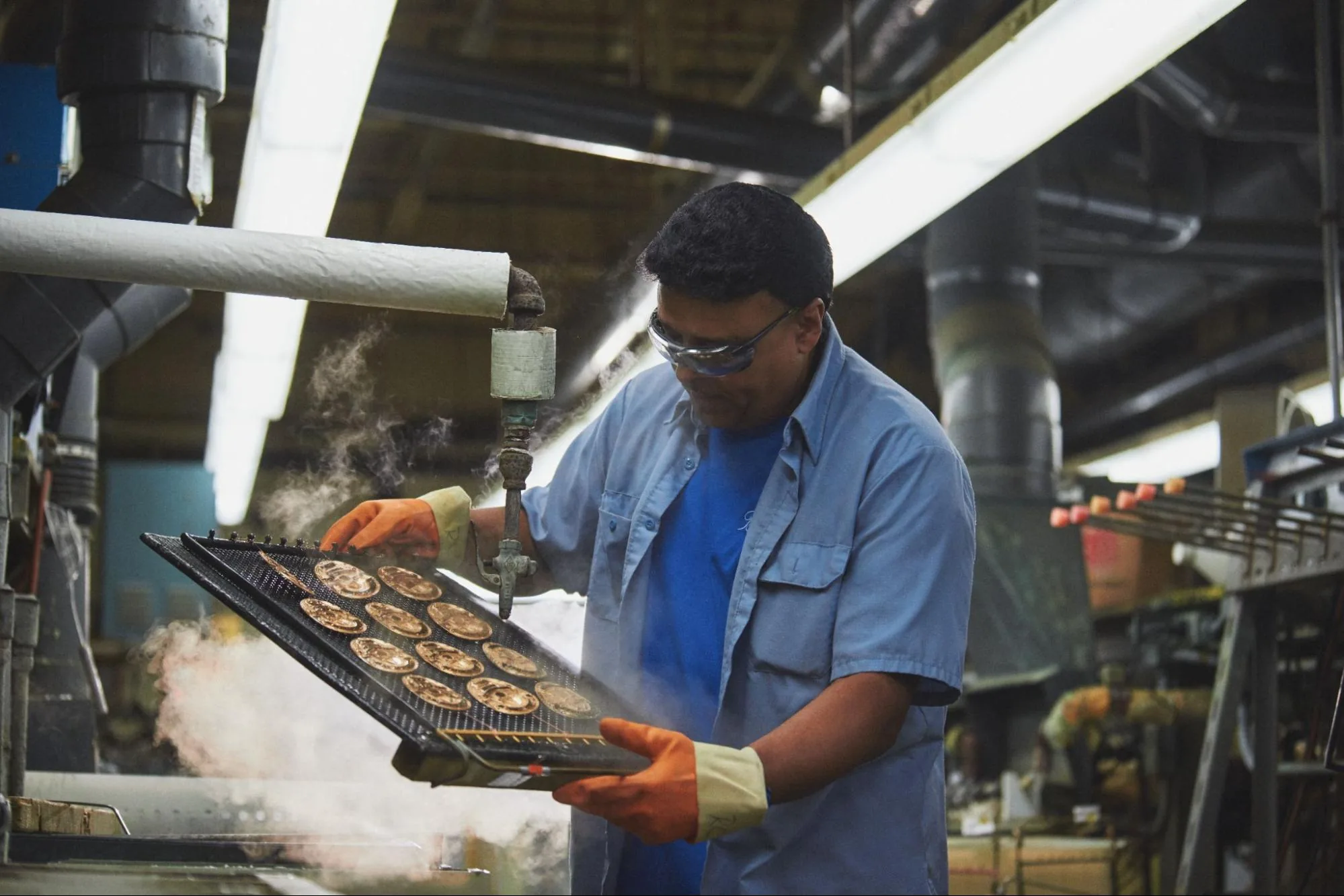 A Blackinton employee in protective gloves steam-cleans a tray of freshly pressed badge medallions on the factory floor.